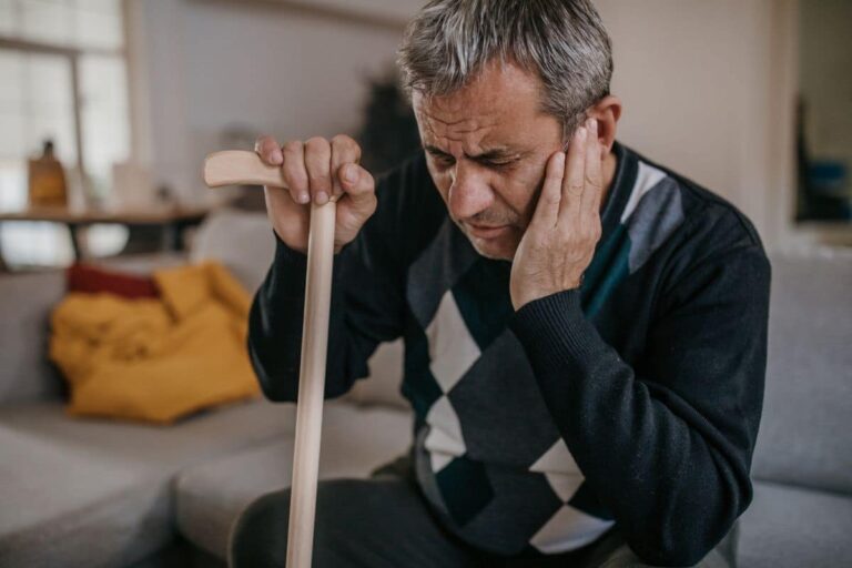 Cholesteatoma Symptoms Older man sitting on the couch and holding himself upright with his cane while holding his ear due to pain from a cholesteatoma